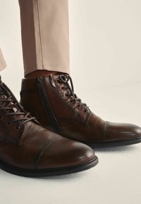 Brązowe skórzane botki za kostkę z okrągłymi noskami, z sznurowaniem i zamkiem błyskawicznym, o gładkiej fakturze i subtelnych detalach w postaci szwów.