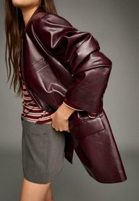 Giacca in pelle bordeaux con maniche oversize, tasca laterale e texture liscia, abbinata a una gonna grigia e una maglietta a righe.