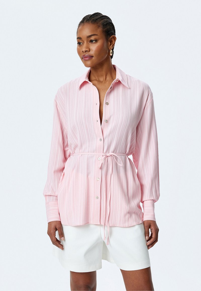 Camisa de botones rosa claro con rayas verticales, mangas largas y cintura ajustable con cordón. Presenta botones de perlas y un corte relajado.