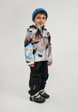Veste d'hiver colorée et à motifs avec des montagnes et des arbres. Pantalons noirs, chaussures grises et beiges. Bonnet en tricot bleu. Enfant souriant.