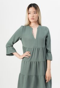Robe verte à volants, avec un décolleté en V, des manches trois-quarts et des poignets froncés, fabriquée à partir d'un tissu léger et texturé.