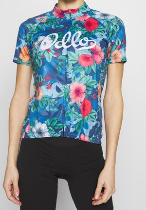 Kvinde iført en blå cykeltrøje med korte ærmer med flerfarvet blomstermønster og hvid tekst, kombineret med sorte cykelshorts.