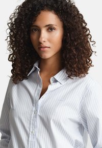 Light blue and white striped button-down shirt with a classic collar, soft cotton fabric, and subtle logo on the chest.