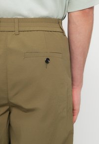 Πλάγια όψη ενός ατόμου που φοράει χακί παντελόνι με κουμπωμένη τσέπη με φάσα και ένα ανοιχτό πράσινο πουκάμισο μέσα στη μέση.