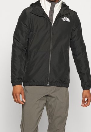 Homme portant une veste North Face noire zippée avec poignets élastiques et capuche, chemise grise boutonnée et pantalon beige.
