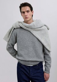 Suéter de punto gris con un dobladillo acanalado, que presenta una tela texturizada. Una bufanda gris más claro está caída sobre los hombros. Jeans de mezclilla oscura.