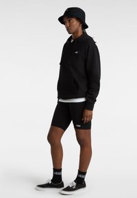 Sudadera negra con bolsillo frontal, combinada con pantalones cortos de ciclismo negros. Un sombrero de cubo negro y zapatillas Vans con acentos blancos completan el look.