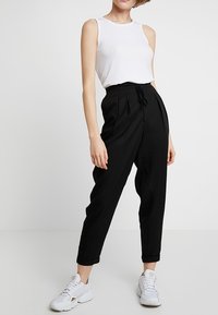 Černé skladané kalhoty s elastickým pasem, kombinované s bílým topem bez rukávů a bílými sportovními botami. Hladká tkanina, volný střih.