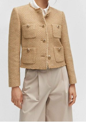 Femme portant une veste en tweed texturé beige avec des boutons dorés et quatre poches frontales, assortie à un pantalon taille haute beige clair.
