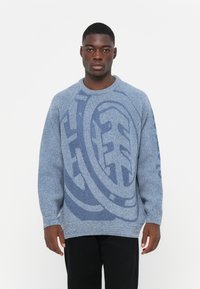 Maglione lavorato a maglia blu con un motivo testurizzato in un blu più scuro. Presenta un collo a giro e polsini a costine, indossato sopra dei pantaloni neri.
