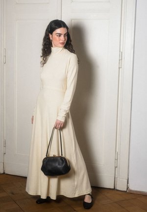 Kvinde med krøllet mørkt hår iført en lang cremefarvet rullekraveskjole, der holder en sort håndtaske og står foran hvide døre.