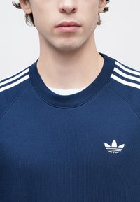 Μπλε σκούρο φούτερ με λευκές τρεις ρίγες στους ώμους και μικρό λευκό λογότυπο. Διαθέτει ριμπ λαιμόκοψη και απαλή υφή.