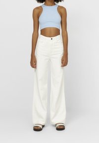 Jean blanc à jambes larges avec taille haute et poches avant, associé à un débardeur court bleu clair. Le modèle porte des sandales plates.