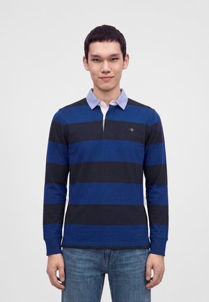Jeune homme portant un polo à manches longues rayé bleu et noir avec un col bleu clair et un jean bleu, debout devant un fond uni.