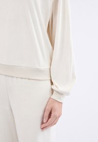 Outfit in tessuto morbido di colore crema, con vita a costine e maniche morbide. Texture liscia, visto di lato con una mano visibile.