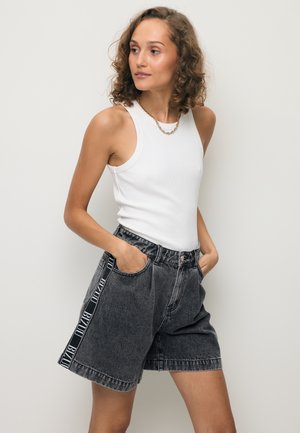 Vrouw met krullend haar die een wit mouwloos topje en zwarte high-waist spijkershort met zijstrepen draagt, staand met handen in de zakken.
