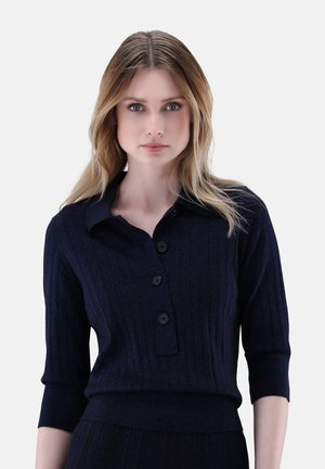 Blonde Frau mit blauen Augen, die einen dunkelblauen, gerippten Pullover mit drei schwarzen Knöpfen und Dreiviertelärmeln trägt.