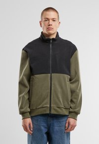 Fleece jacket featuring a zip front, high collar, color-block design in black and olive green, with side pockets and a soft texture.