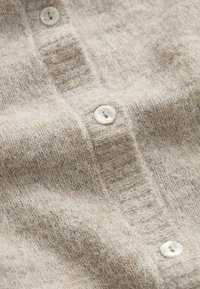 Beige gebreide cardigan met een zachte, gestructureerde oppervlakte en kleine, ronde, witte knopen. Vert toont een fijn steekpatroon en een lichte fuzzy uitstraling.
