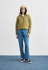 Πράσινο fleece πουλόβερ με τετράγωνο φερμουάρ και μπροστινές τσέπες, συνδυασμένο με μπλε τζιν παντελόνι με φαρδιά πόδια και μαύρα αθλητικά παπούτσια. Ελαφρύ καπέλο ολοκληρώνει την εμφάνιση.