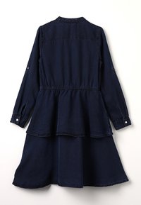 Tummansininen pitkähihaisessa mekossa, jossa on napitettu hihansuu, kerätty vyötärö ja kaksitasoinen helma, kuvattu takaa valkoista taustaa vasten.