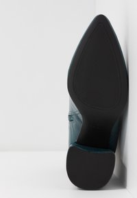 Semelle d'une botte à bout pointu avec talon et avant-pied en caoutchouc noir texturé, sur un fond de mur et de sol blancs.