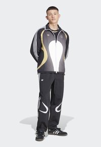 Jauns vīrietis stāv ar rokām aiz muguras, ģērbies melnā un zelta Adidas treniņjakā, melnās biksēs ar baltām svītrām un melnās sporta apavās.