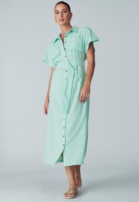 Vestito verde menta con bottoni, maniche corte, due tasche frontali e una cintura abbinata. Presenta una silhouette dritta e un'orlo a lunghezza ginocchio.