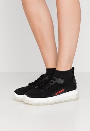 Osoba nosząca czarne buty sportowe Love Moschino w stylu skarpetek z białymi grubymi podeszwami, stojąca na jednolitej jasnoszarej podłodze.