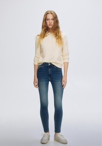 Donna con capelli lunghi e mossi che indossa un maglione polo beige a maniche lunghe, jeans skinny blu a vita alta e sneakers bianche, in piedi davanti a uno sfondo semplice.