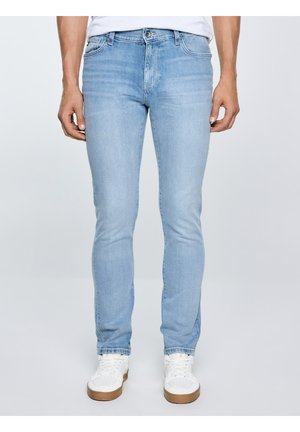 Mees, kes kannab helehalli kitsaid teksapükse ja valgeid tenniseid, seisab ühevärvilise taustaga.