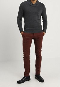 Uomo che indossa un maglione a maglia grigio scuro con colletto bottonato, pantaloni attillati color ruggine e stivali Chelsea in pelle nera, con le mani in tasca.