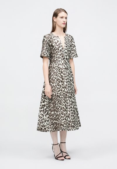 Γυναίκα που στέκεται, φοράει ένα μεσομήκος φόρεμα με λεοπάρ σχέδιο και κοντά μανίκια, συνδυασμένο με λευκά καλσόν με σχέδια και μαύρα πέδιλα με λουράκια.