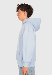 Junger Mann mit lockigem Haar, der einen hellblauen Kapuzenpullover und hellblaue Jeans trägt, und im Profil vor einem weißen Hintergrund steht.