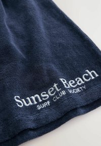 Dunkelblaues Handtuch aus Frottee mit weicher Textur, mit weißem gesticktem Text "Sunset Beach Surf Club Society" am Rand.