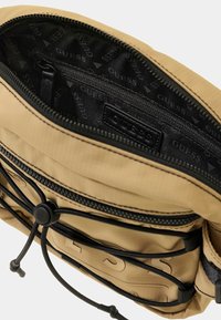 Beige Nylon Tasche mit Reißverschluss, Innentaschen und schwarzen Kordel-Akzenten. Abgefüttert mit einem sich wiederholenden Logomuster.