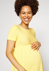 Vestido de maternidade amarelo com mangas curtas e um corpete texturizado e plissado. A saia é solta e fluida, projetada para conforto.
