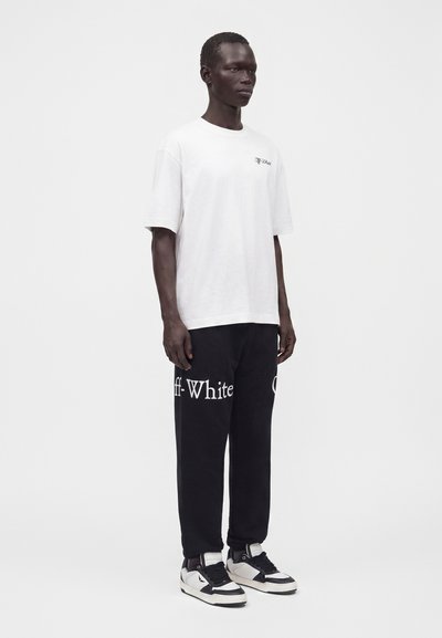 Homme debout portant un t-shirt blanc à manches courtes, un pantalon de survêtement noir avec un grand texte blanc "Off-White", et des baskets noires et blanches.