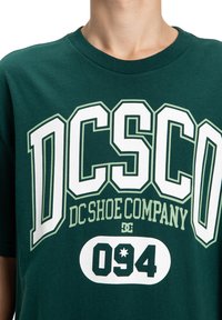 Zielona bawełniana koszulka z dużym białym i kremowym napisem graficznym "DC Shoe Company" oraz liczbą "094" w odważnej, sportowej czcionce.
