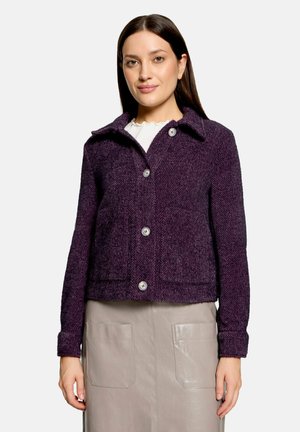Femme aux longs cheveux foncés portant une veste en laine violet foncé boutonnée sur un haut blanc et une jupe beige avec des poches plaquées à l'avant.