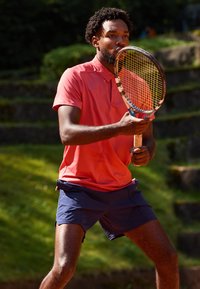 En spiller i en lys korallpolo skjorte og marineblå shorts holder en tennisracket. Området har grønt gress og steintrapper i bakgrunnen.