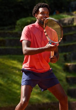 Ein Spieler in einem hellen Korallen-Polohemd und navyblauen Shorts hält einen Tennisschläger. Im Hintergrund sind grünes Gras und steinerne Stufen zu sehen.