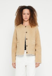 Beżowa kurtka z okrągłym dekoltem, zapinana na guziki, z dwoma przednimi kieszeniami i kontrastującymi przeszyciami, wykonana z miękkiego materiału, przedstawiona na neutralnym tle.