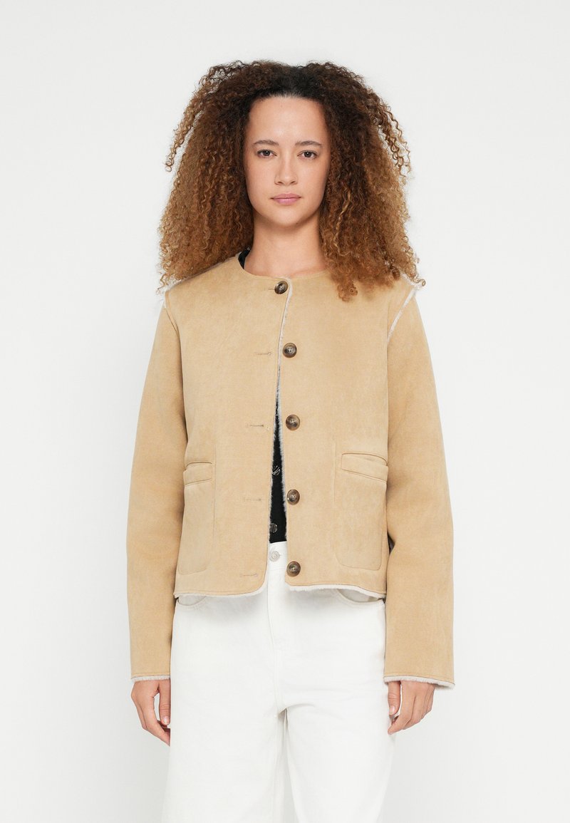 Beżowa kurtka z okrągłym dekoltem, zapinana na guziki, z dwoma przednimi kieszeniami i kontrastującymi przeszyciami, wykonana z miękkiego materiału, przedstawiona na neutralnym tle.