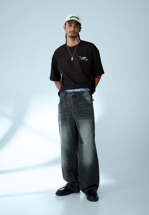 Moški, ki nosi črno preveliko majico z napisom "Nothing", izprane široke kavbojke, črne čevlje, belo kapo in ogrlico z obeskom, stoji sproščeno.