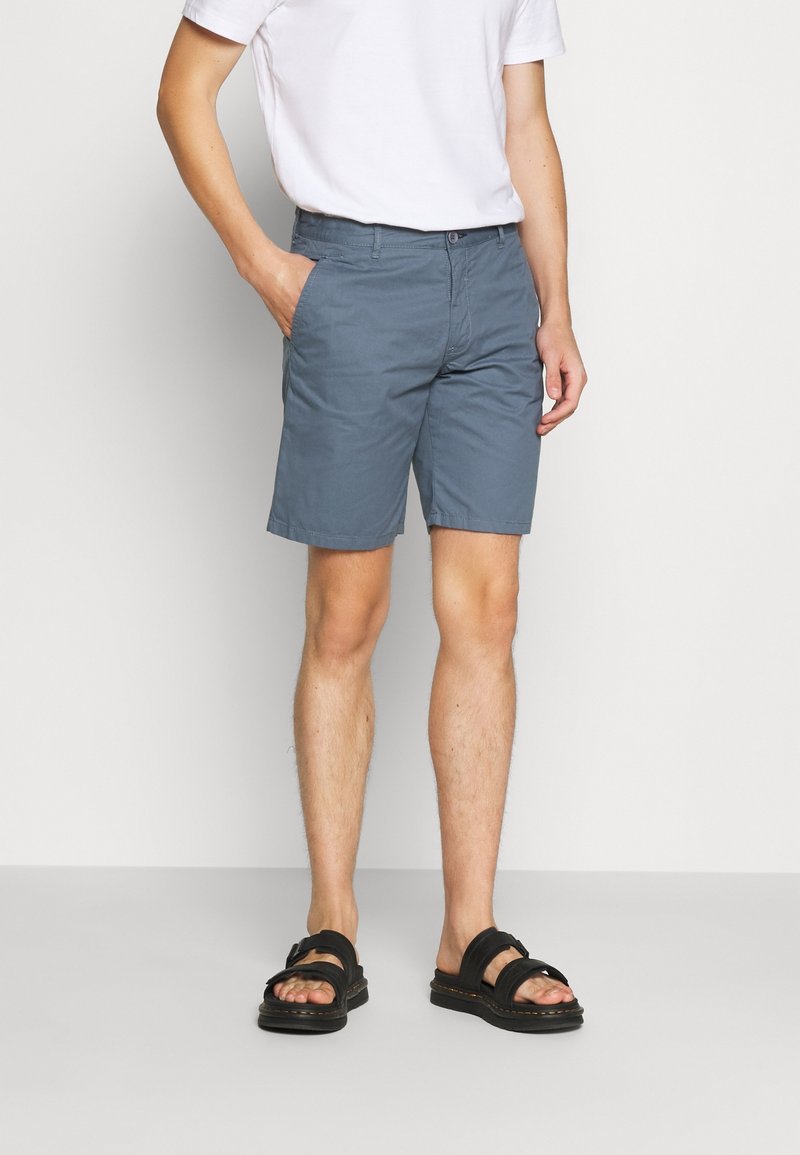 Pantalones cortos de algodón gris para hombre con corte recto, con bolsillos laterales y cierre de botón. Combinados con sandalias negras de puntera abierta.