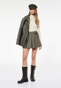 Grau kariertes Sakko über einem cremefarbenen Rollkragenpullover, kombiniert mit einem grauen Faltenrock mit diagonalem Riemen und braunen knielangen Stiefeln.