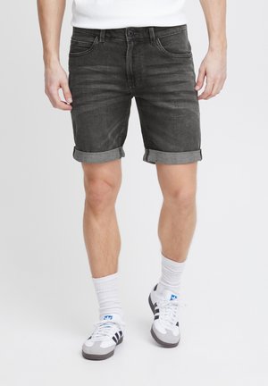 Homme portant un short en denim noir retroussé, des chaussettes blanches et des baskets Adidas blanches à rayures noires, debout devant un fond uni.