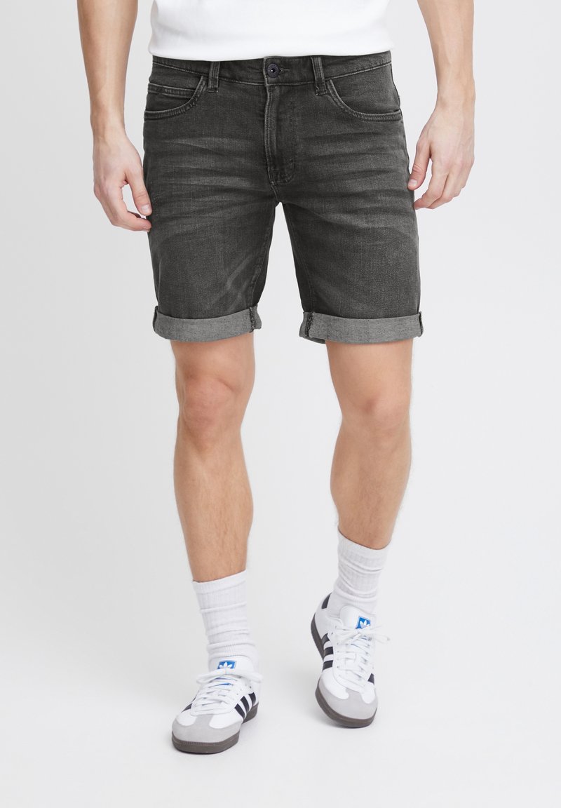 Homme portant un short en denim noir retroussé, des chaussettes blanches et des baskets Adidas blanches à rayures noires, debout devant un fond uni.