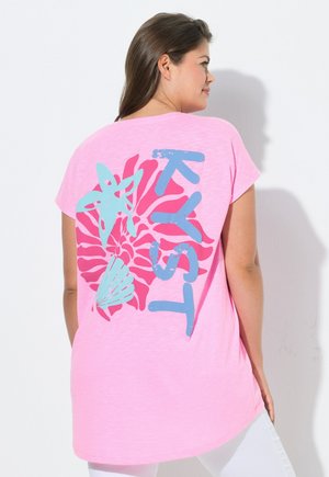 Vrouw in een roze T-shirt met blauw en turquoise zeesterren-, schelp- en spiraalgrafisch ontwerp en gestileerde tekst op de achterkant, draagt een witte broek.